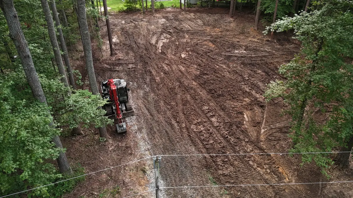 Overhead view of a cleared, track-marked lot in the woods, with a red excavator on a gravel entrance