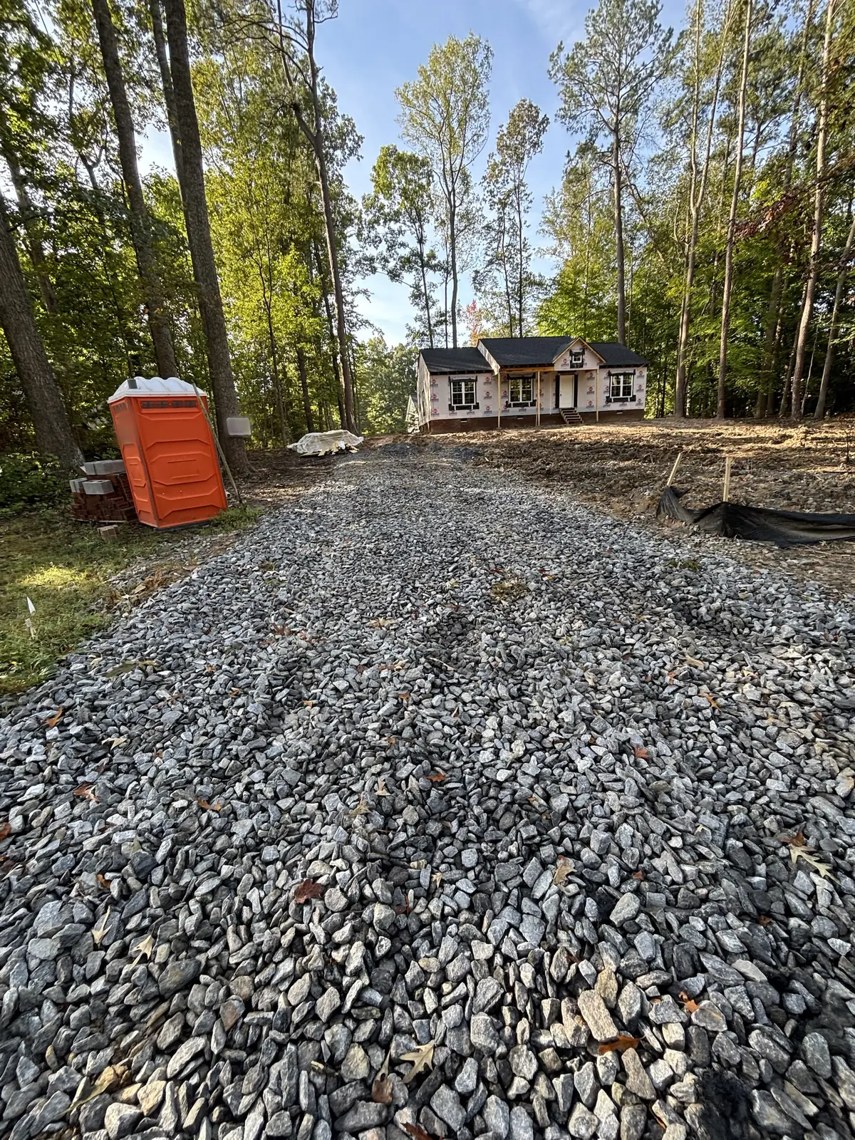 New gravel driveway leading to a house under construction among tall trees, with a portable toilet and building materials beside it