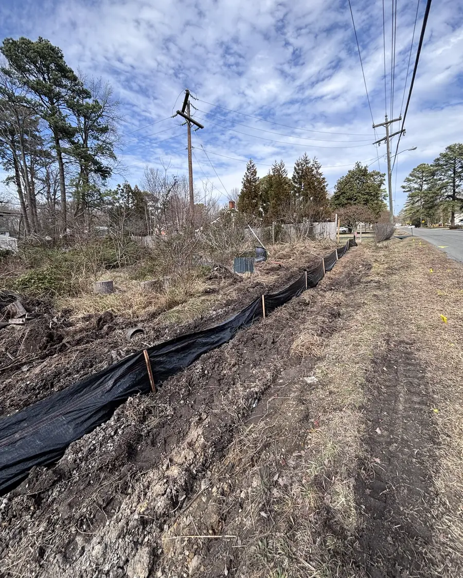 Black silt fence on wooden stakes along freshly cleared ground beside a residential road, with yellow utility-locate flags in the grass and power lines overhead